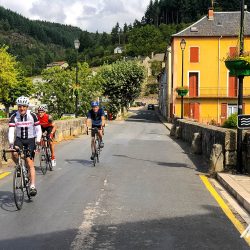 Cyclists crossing bridge over River Dourbie in rural village on Marmot Tours Raid Massif Central French cycling holidays