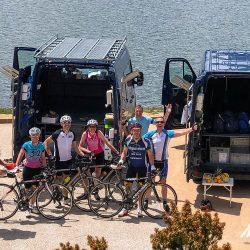 Group of cyclists waving and enjoying snacks offered by Marmot Tours guide at support vehicle on coast road of Raid Corsica