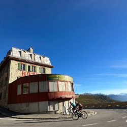 Pair of cyclists riding past Hotel Belvedere on Furka Pass cycling climb Swiss Alps on Marmot Tours Raid Dolomites challenge