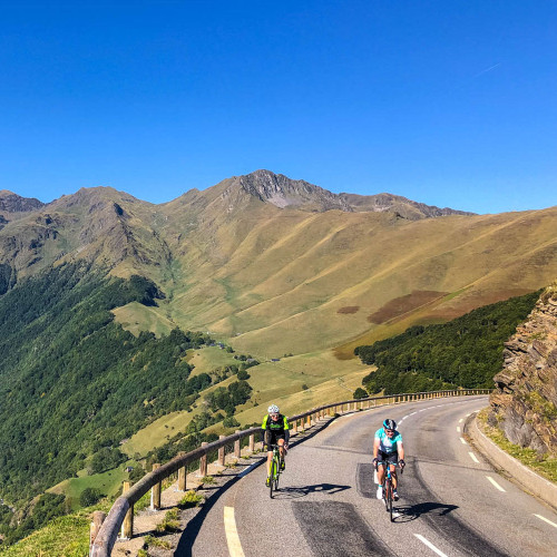 pair of cyclists climbing Col de Peyresourde on marmot tours road cycling holiday in French Pyrenees with green mountain summits in background and bright blue sky