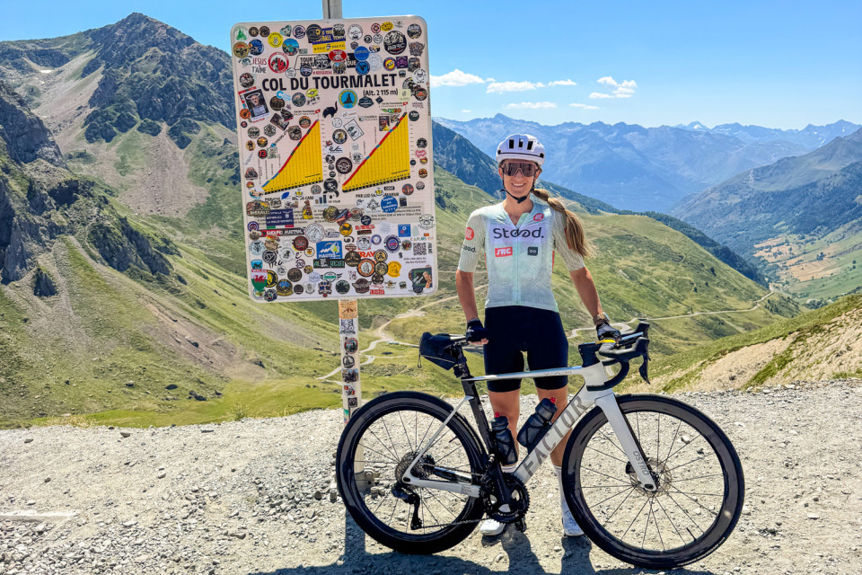 female cyclist posing by col du Tourmalet sign with pyrenean peaks backdrop on marmot tours guided road cycling holiday French Pyrénées