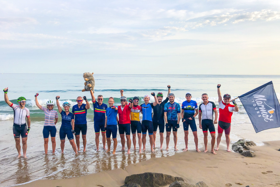 group of cyclists pose for photo with marmot tours flag on beach at Atlantic coastline on marmot tours Atlantic to mediterranean cycling challenge