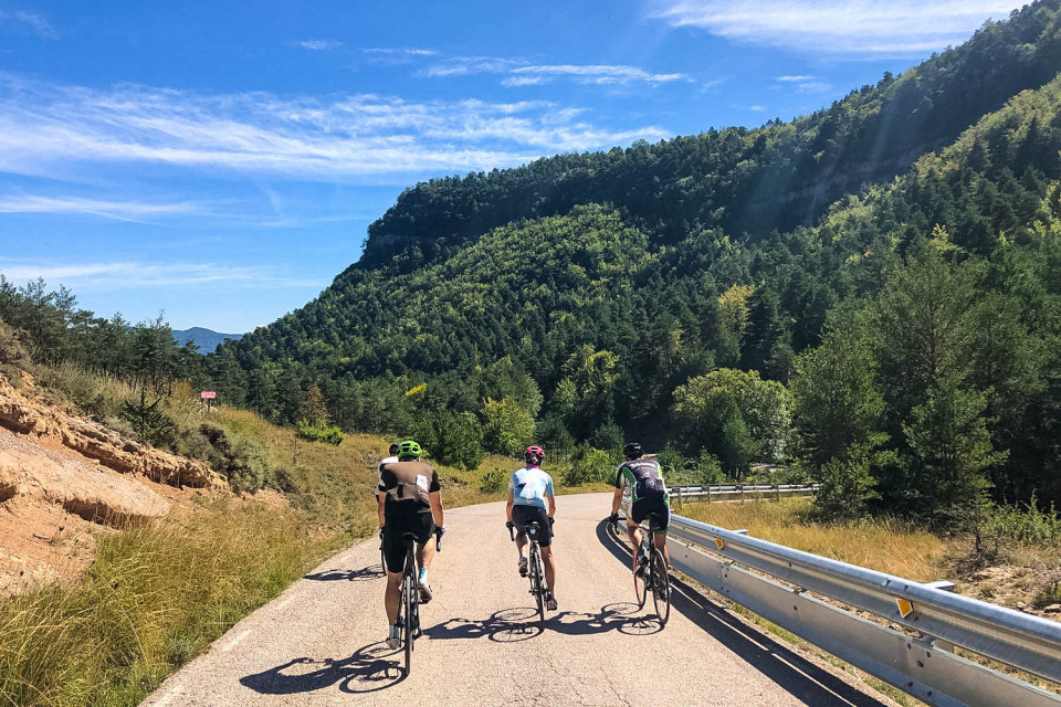 Three cyclists pedal away towards forest under blue sky on guided road cycling tour Catalonia with Marmot Tours