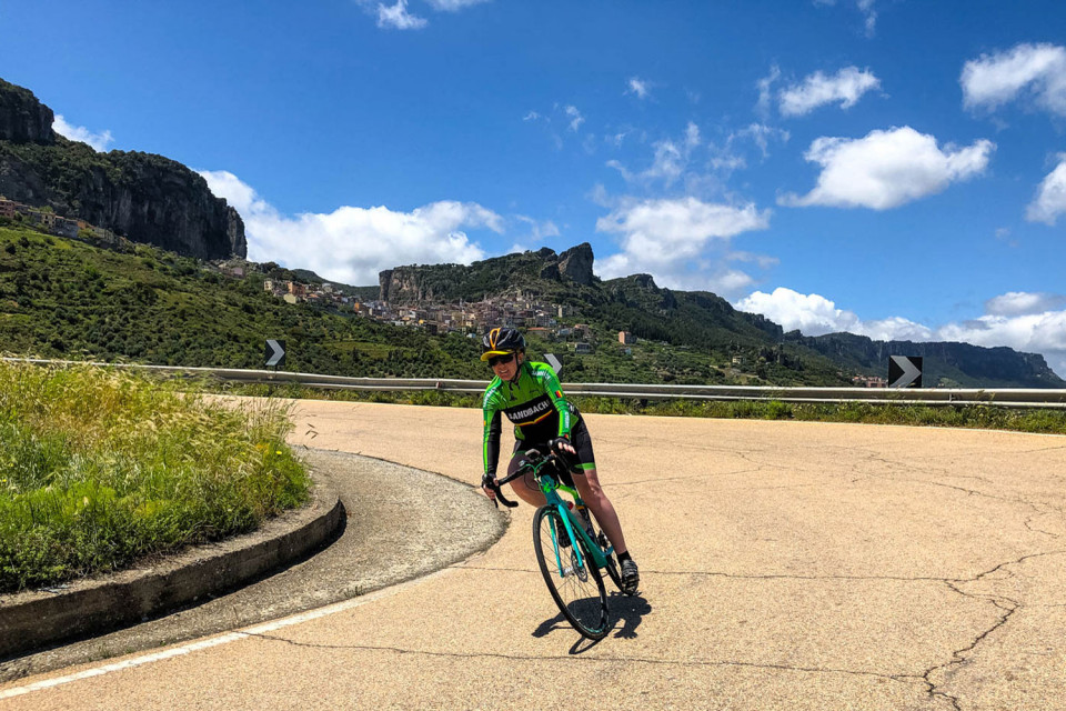 Female cyclist descending hairpin bend under blue sky with escarpment backdrop on guided road cycling tour of Sardinia with Marmot Tours