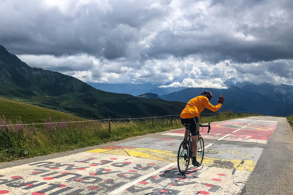 Cyclist riding away waving on Madeline cycling climb with graffiti on road and alpine peaks on Marmot Tours guided road cycling mini break French Alps