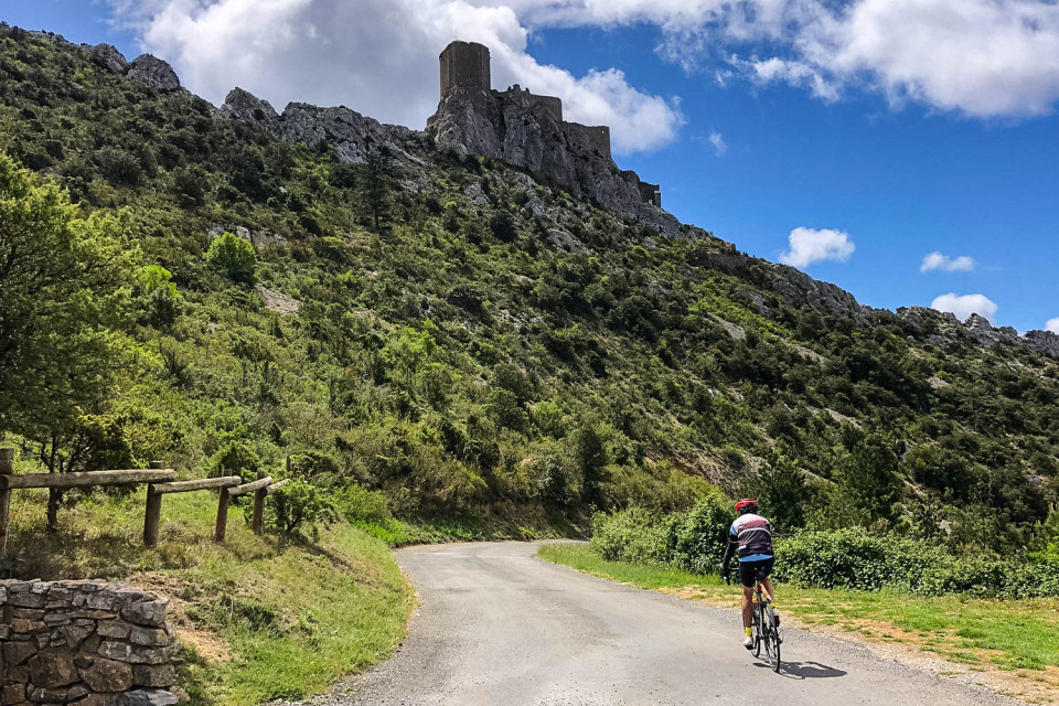 Cyclist climbing road beneath chateaux ruins on Marmot Tours guided road cycling tour Foothills of Pyrenees