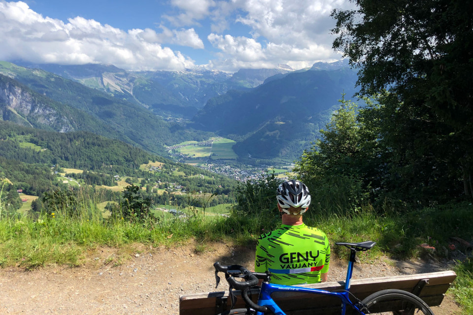 Cyclist seated on bench admiring Col de Joux Plane climb with Alpine valley and peaks on Marmot Tours guided road cycling tour Northern French Alps