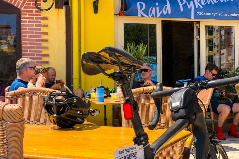 group of cyclists celebrate at outside bar terrace with bike in foreground at finish of raid pyrenees with marmot tours