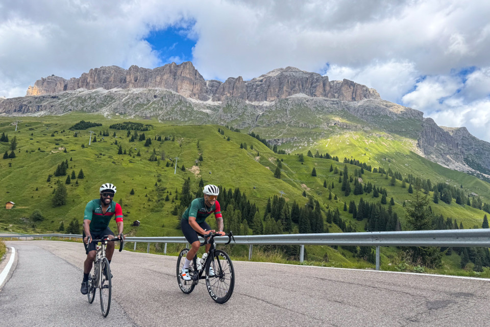 pair of cyclists in marching green and red kit climb passo pordoi with dolomites peaks backdrop on marmot tours cycling mini break