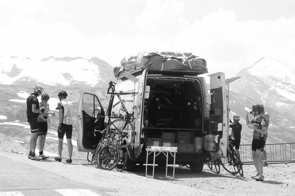 Black and white image of cyclists enjoying snacks from Marmot Tours support vehicle on Raid Alpine