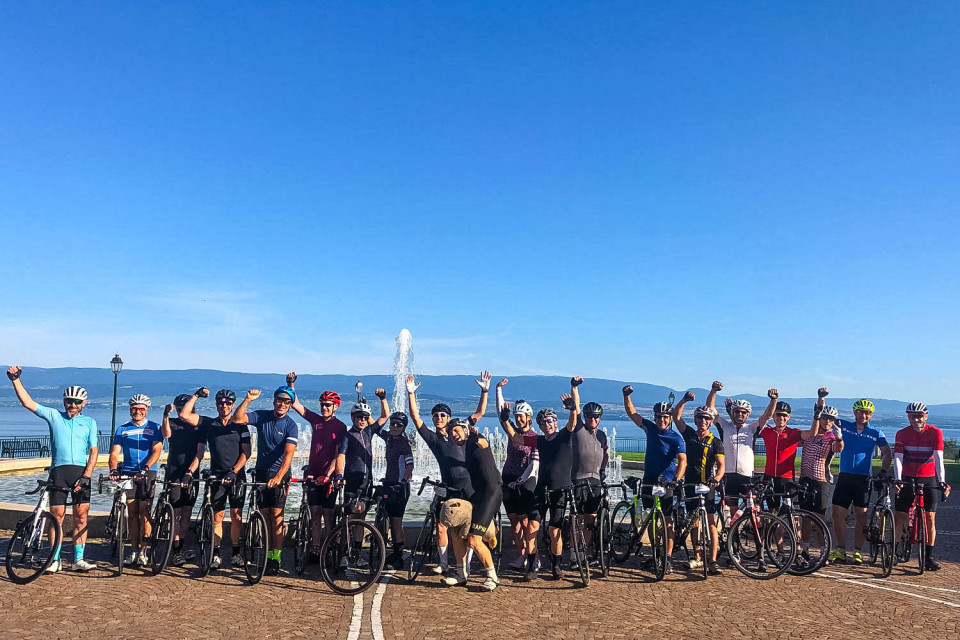 Group of cyclists pose on shore of lake Geneva at start of raid alpine cycling challenge with marmot tours