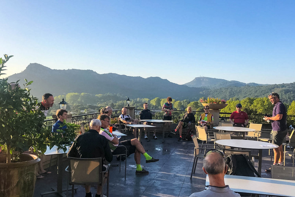 Group of cyclists enjoy drinks and briefing on hotel terrace with mountains backdrop on Marmot Tours guided road cycling holiday