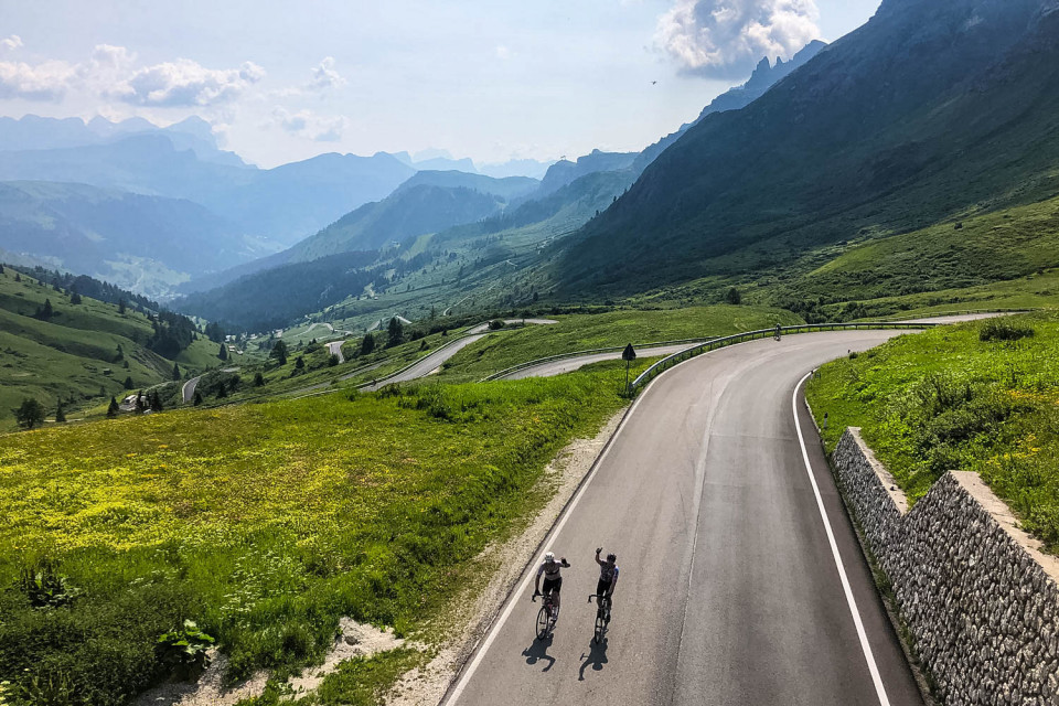 Pair of cyclists waving before riding under bridge with Italian Dolomites backdrop on Marmot Tours guided cycling holiday Italy