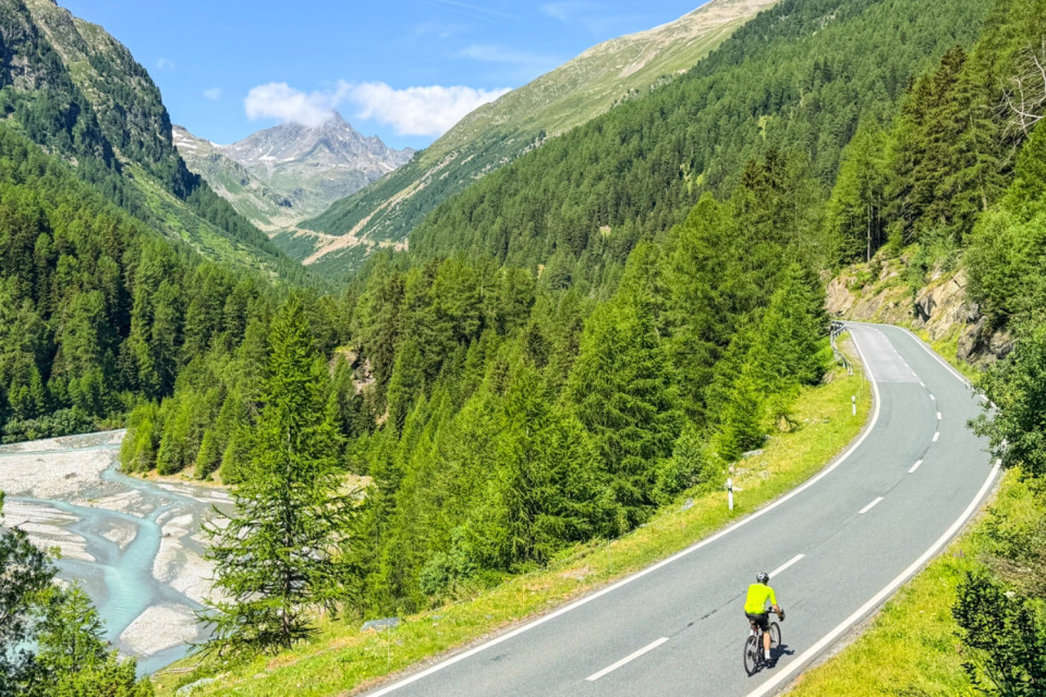 Cyclist climbing pass through valley alongside glacial stream with mountain peaks in background on marmot tours guided road cycling holiday Italy