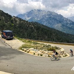 Pair of cyclist rounding hairpin bend with Marmot Tours support vehicle ahead on mountain climb of Raid Corsica