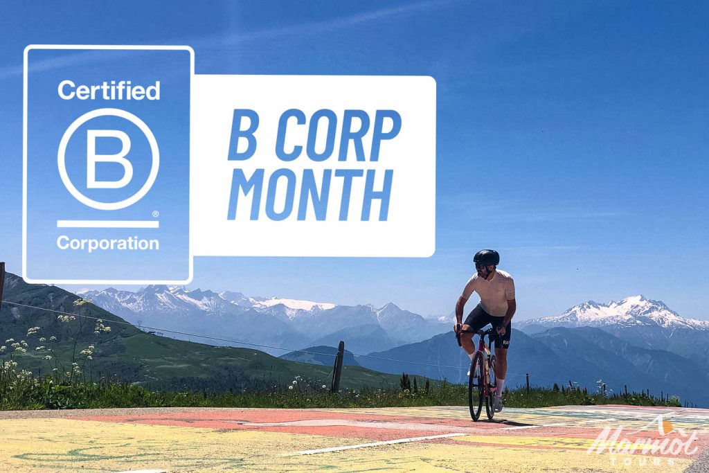 Cyclist climbing Col de la Madeleine French Alps on Marmot Tours road cycling tour beneath blue skies and snowy mountain peaks with B Corp Month logo overlaid