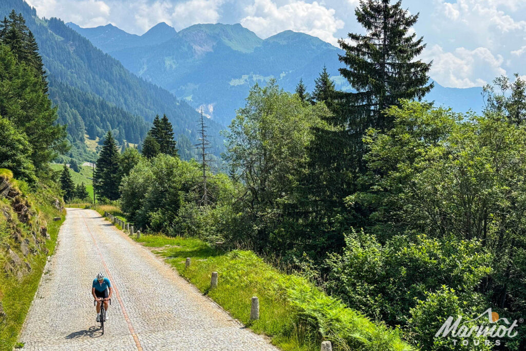 Cyclist climbing St Gotthard pass Tremola road in Swiss Alps with Marmot Tours mountainous backdrop and forests