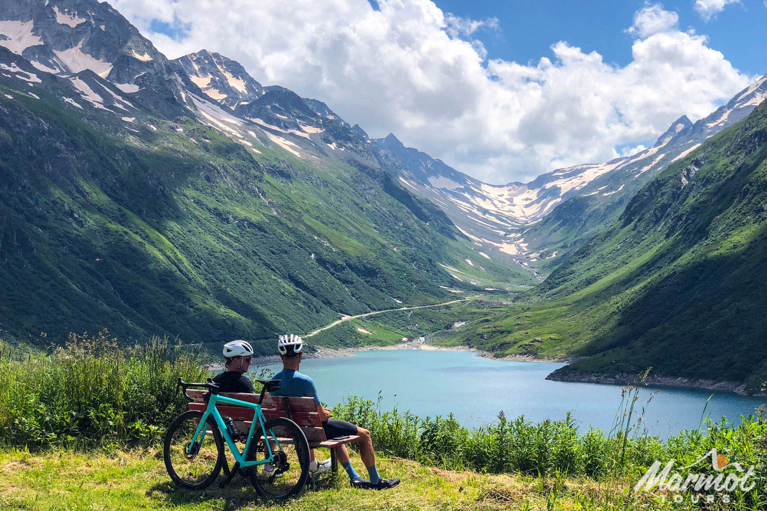 Pair of cyclists sitting on bench in Swiss Alps admiring glacier and lake with Marmot Tours road cycling tours