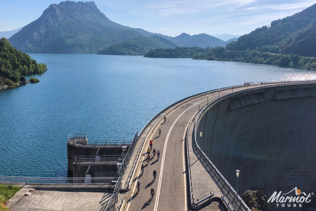 Group of cyclists riding over dam with reservoir and forested mountains backdrop on Marmot Tours guided road cycling tour Picos Spain