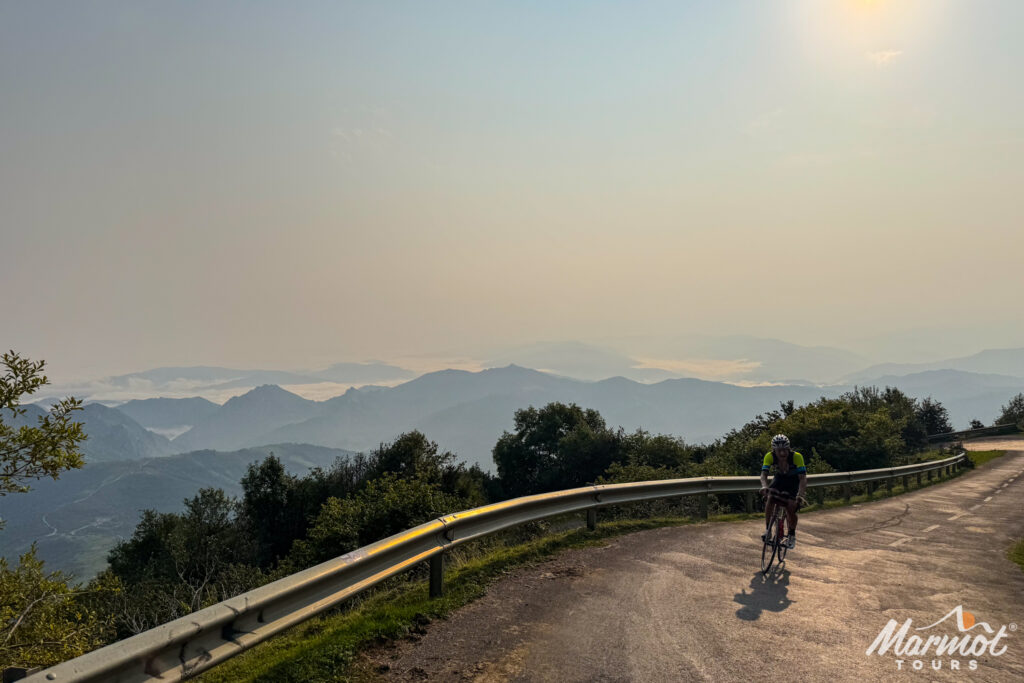 Cyclist climbing Alto de l'Angliru in the Picos of Northern Spain with mountain peaks in distance on Marmot Tours guided road cycling holiday