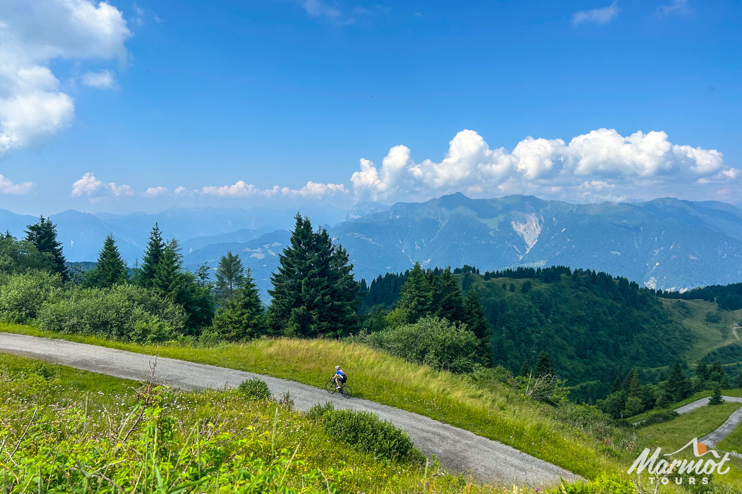 A cyclist nearing summit of Monte Zoncolan with mountain peaks backdrop and blue cloudy sky on Marmot Tours guided road cycling holiday Slovenia Italy Austria