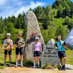 Group of cyclists celebrate at Passo del Mortirolo sign and Marmot Tours flag on guided road cycling holiday Italian Dolomites and Swiss Alps