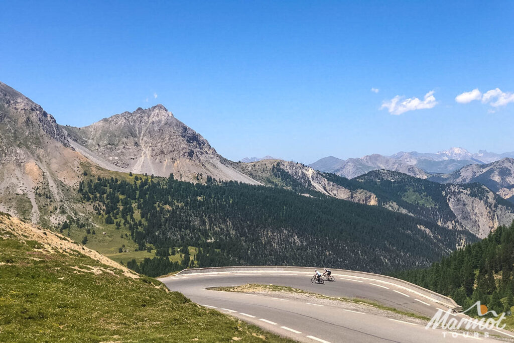 Pair of cyclists approach switchback on Col d'Izoard with mountain peaks backdrop on Raid Alpine cycling challenge with Marmot Tours