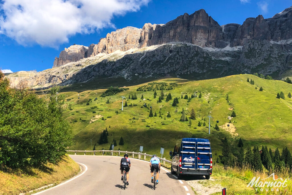 Pair of cyclists climb Passo Pordoi with mountain ridge backdrop passing Marmot Tours support vehicle on guided road cycling tour Italian Dolomites