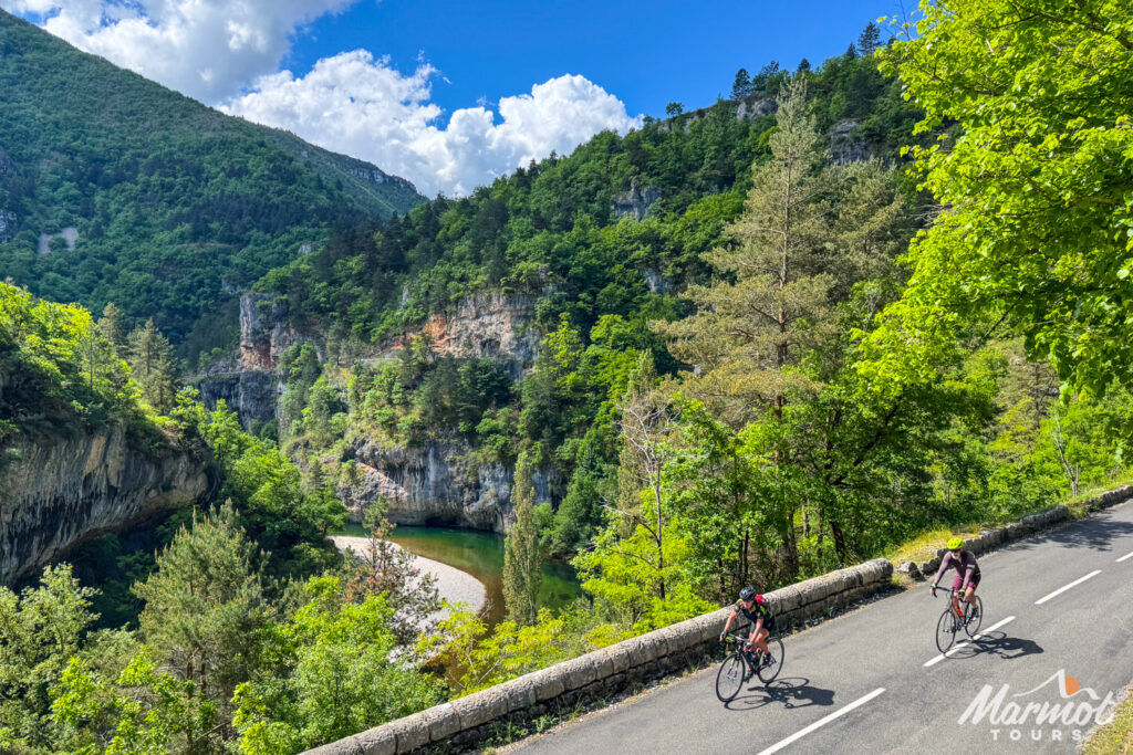 Pair of cyclists riding in Gorge du Tarn surrounded by forested slopes and river Tarn below on guided road cycling holiday Cevennes and Ardeche south of France with Marmot Tours
