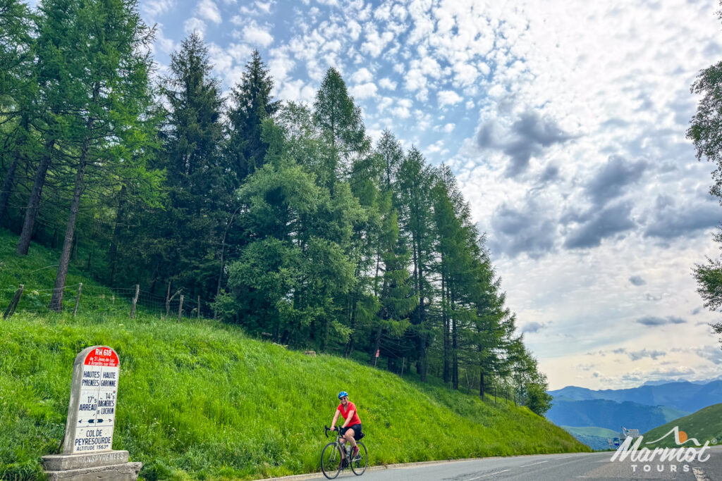 Female cyclist summiting Col de Peyresourde on pine tree lined mountain road under cloudy sky on Marmot Tour cycling holiday in French Pyrenees