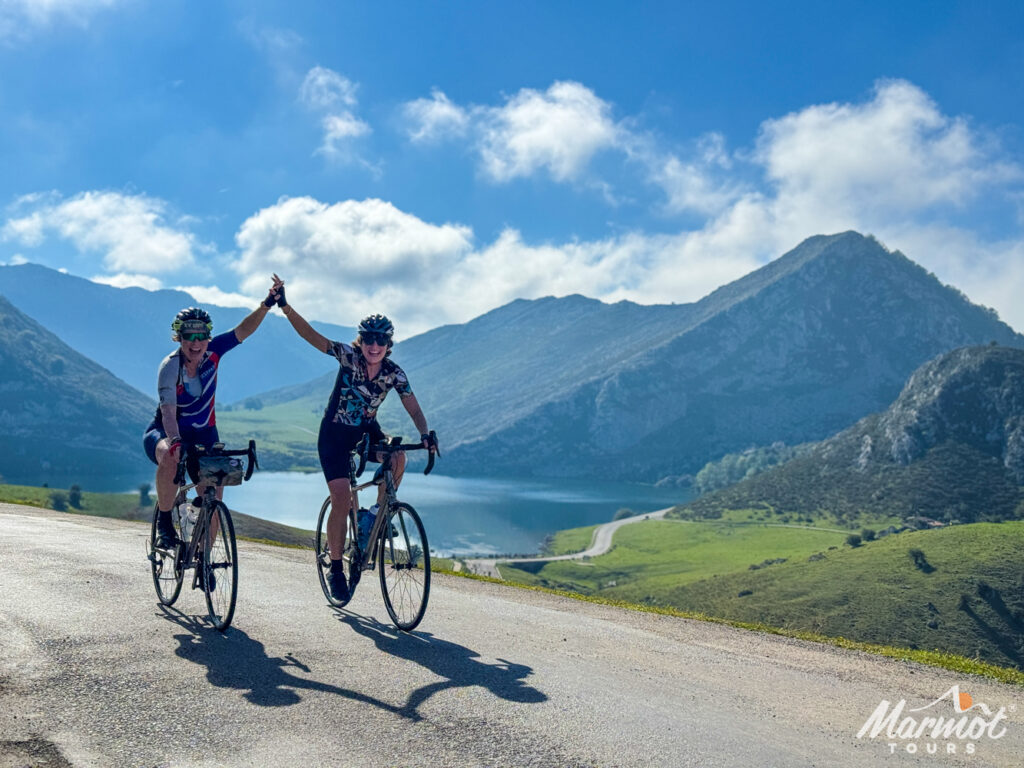 Pair of female cyclists summiting Lagos de Covadonga hand in hand surrounded by lake and mountain peaks on guided cycling holiday in Spain with Marmot Tours