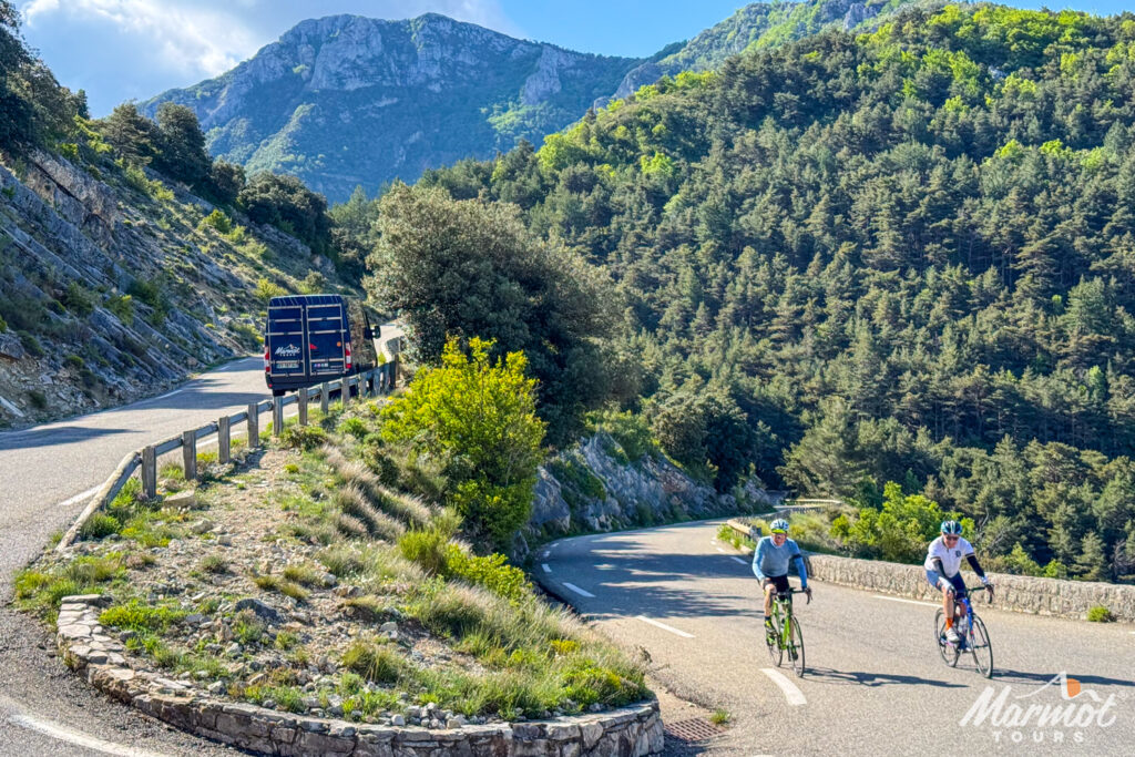 Pair of cyclists approach hairpin bend with Marmot Tours vann ahead surrounded by forested slopes on guided cycling holiday in Provence Ventoux Verdon Gorge