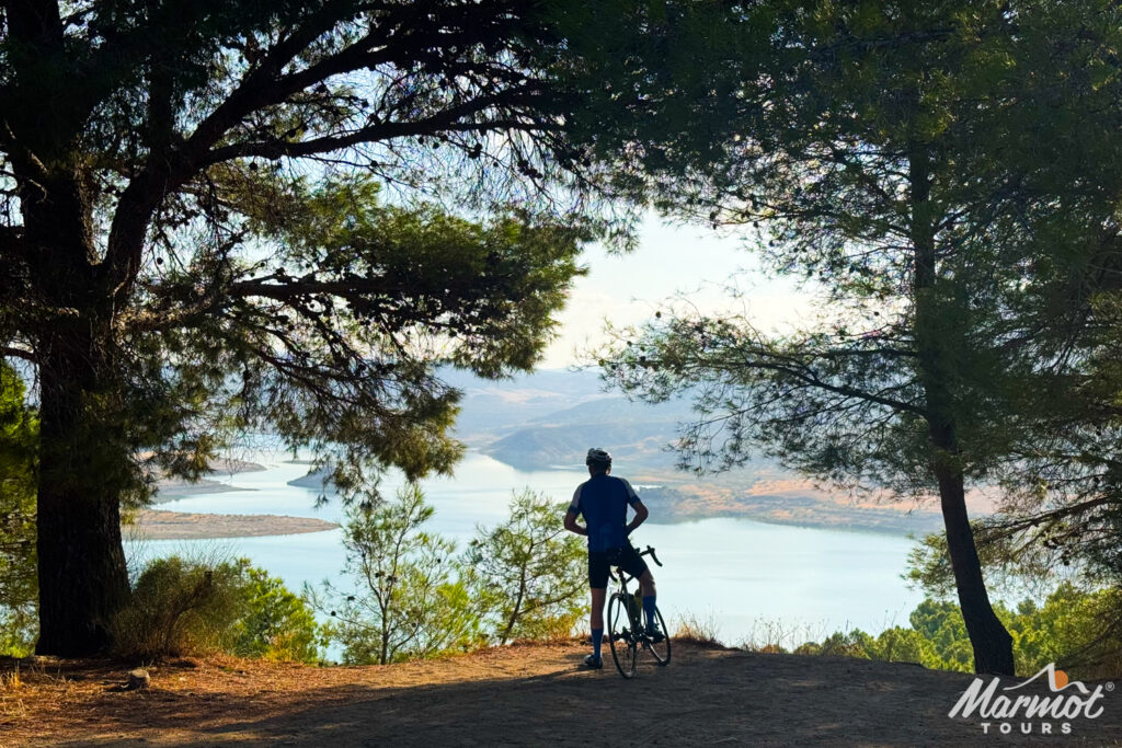 Cyclist pausing to view reservoir beneath canopy of trees on guided road cycling holiday Andalusia with Marmot Tours